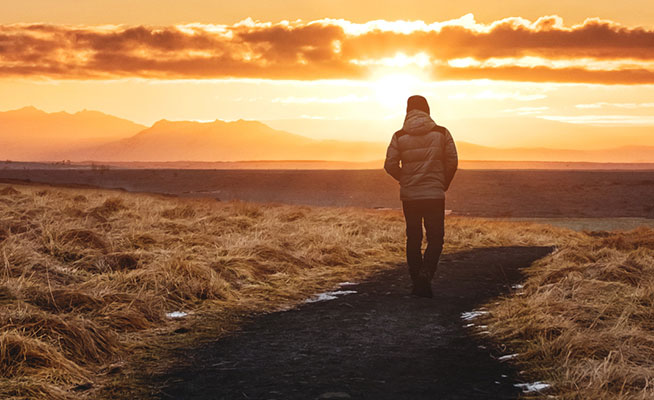 Man walking down path through an open field into the sunset