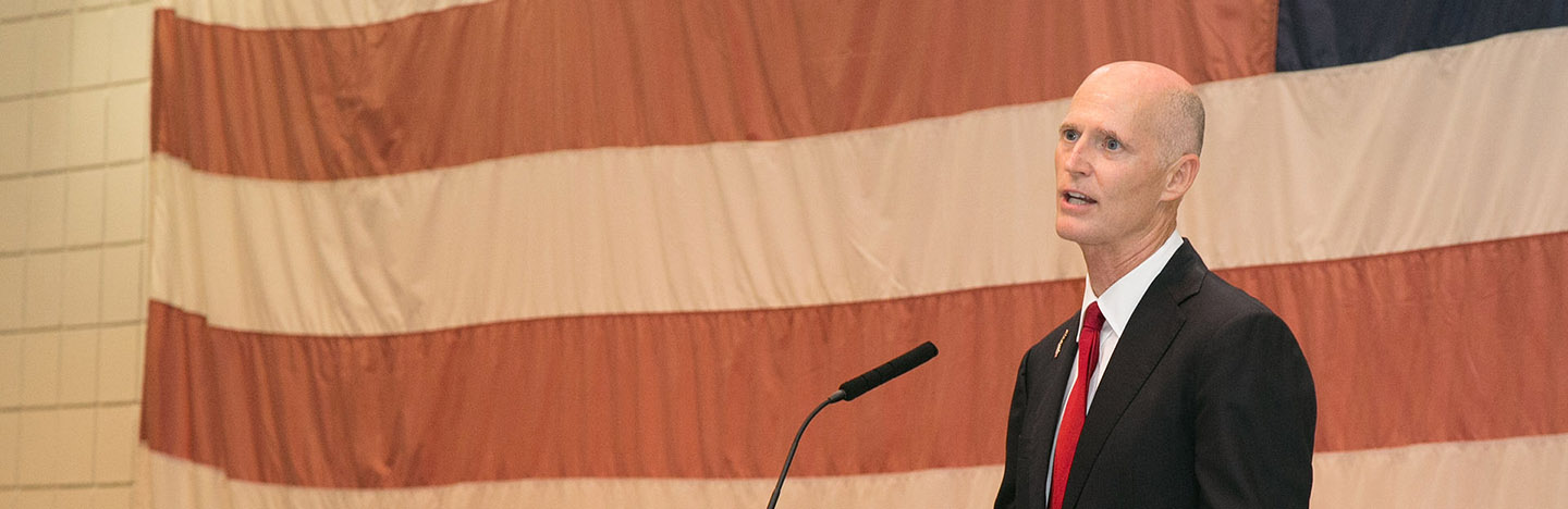 Governor Rick Scott speaking at a podium in front of an American flag